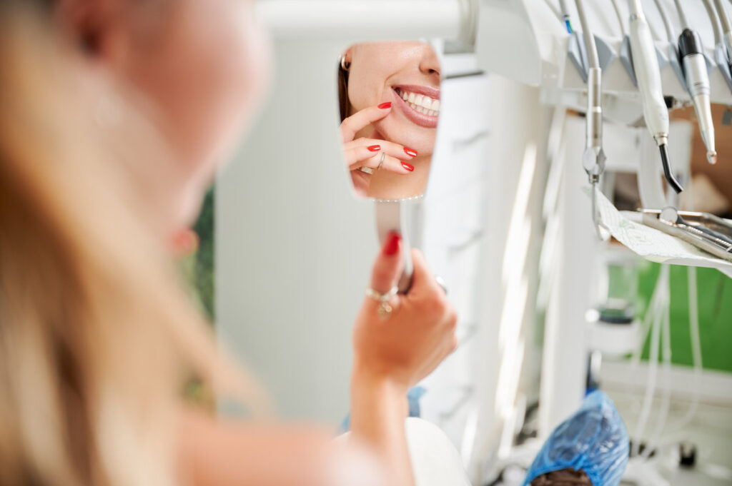 Female patient admires healthy teeth in mirror at dental office. Close up reflection